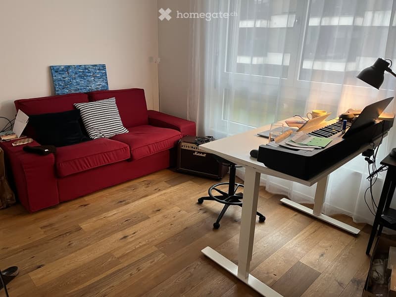 A modern living room with a red sofa, a standing desk with a laptop, and a floor lamp near a large window with sheer curtains.