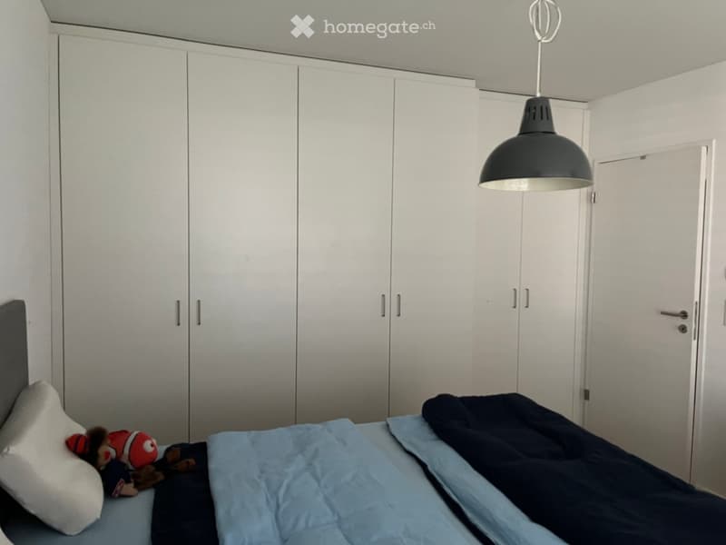 A modern, minimalist bedroom with a large bed, white built-in wardrobes, and a single hanging light fixture. The bed is made with blue and dark blue bedding and features a stuffed toy on the pillow.