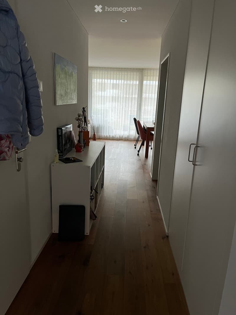 A narrow hallway with wooden flooring, white walls, and a small console table with items on it. A doorway leads to another room with a desk and chairs visible.