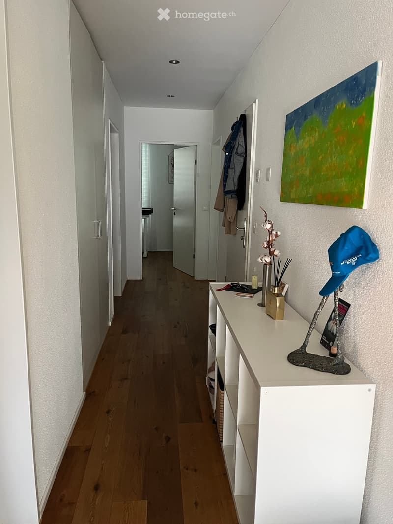 A narrow hallway with wooden flooring, white walls, and a small console table holding decorative items, including a vase with flowers, a sculpture, and a hat.