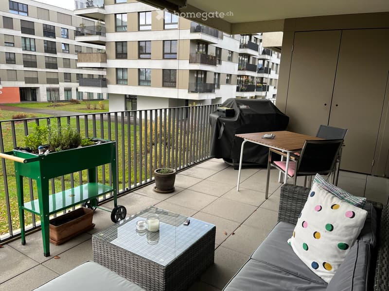 Balcony with a sofa, coffee table, dining table, chairs, a bar cart, and a barbecue, overlooking a modern residential complex.