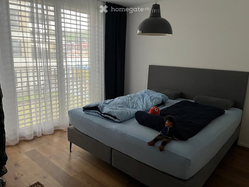 A modern bedroom with a large bed, headboard, pillows, and a blanket. A toy and a plush toy are placed on the bed. The room features wooden flooring and large windows with sheer curtains.