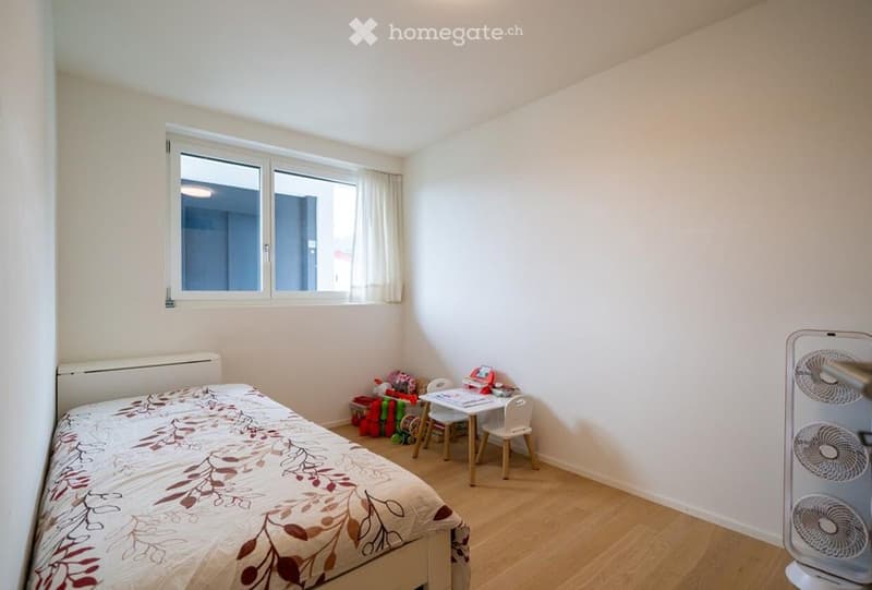 A small, well-lit bedroom with a single bed, a small table and chair, and a fan near the window. The room has light wooden flooring and white walls.