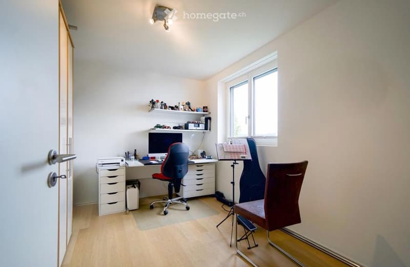 A well-lit room with a wooden floor, a desk with a computer and chair, a guitar on a stand, a shelf with various items, and a printer on a cabinet.