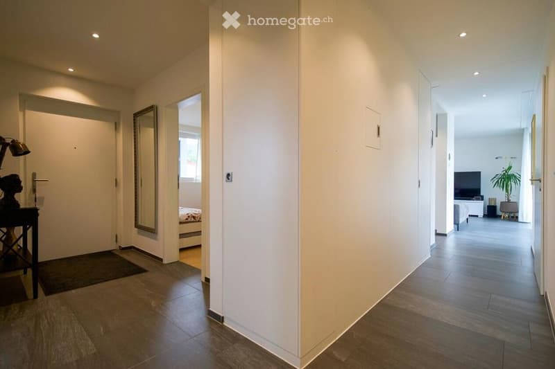 Modern hallway with wooden flooring, white walls, recessed lighting, and a small table with a decorative item on the left side. The hallway leads to a bedroom and a living area with a TV and seating.