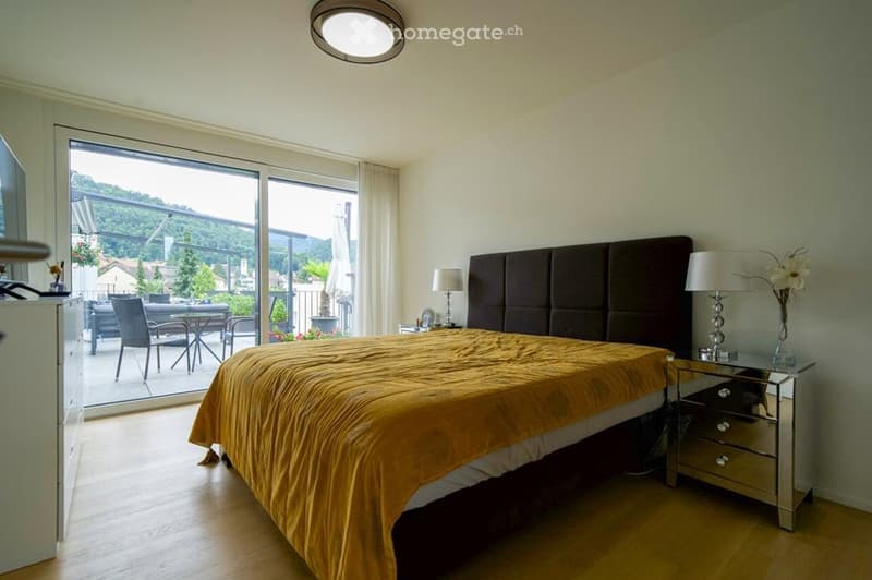 Modern bedroom with large windows offering a scenic view, featuring a large bed with a dark headboard, a nightstand with a lamp, and a small table with chairs visible through the window.