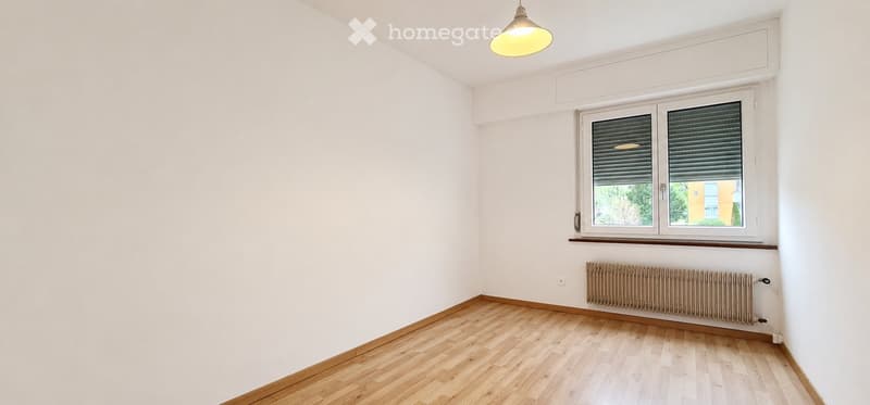 Empty room with light wooden flooring, white walls, a single window with blinds, a radiator, and a hanging light fixture.