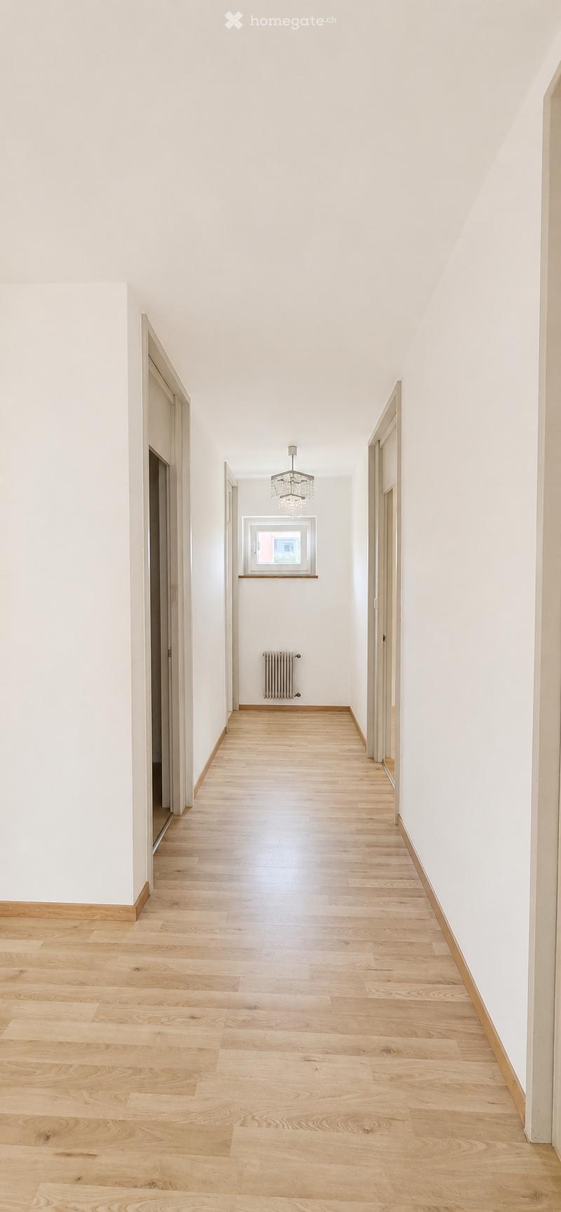 Bright, minimalist hallway with light wooden flooring, white walls, and a chandelier hanging from the ceiling. Features a radiator and a window at the far end.