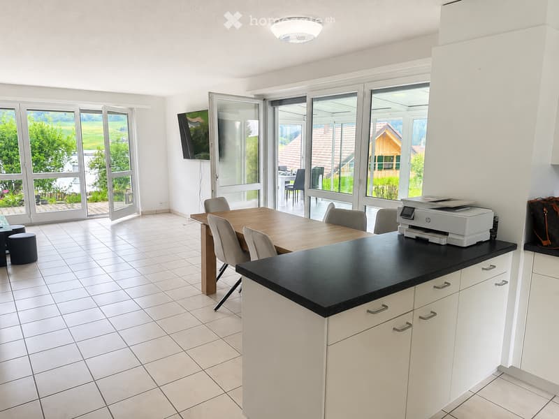 Bright kitchen with white cabinets, black countertop, tiled floor, and large windows overlooking a green landscape. Includes a dining table with chairs, a printer on the counter, and a TV mounted on the wall.