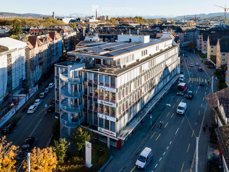 multi-story commercial building with solar panels on the roof, multiple street-facing windows, ground-level businesses, spiral staircase at the back, parking spaces on the street, surrounding urban environment