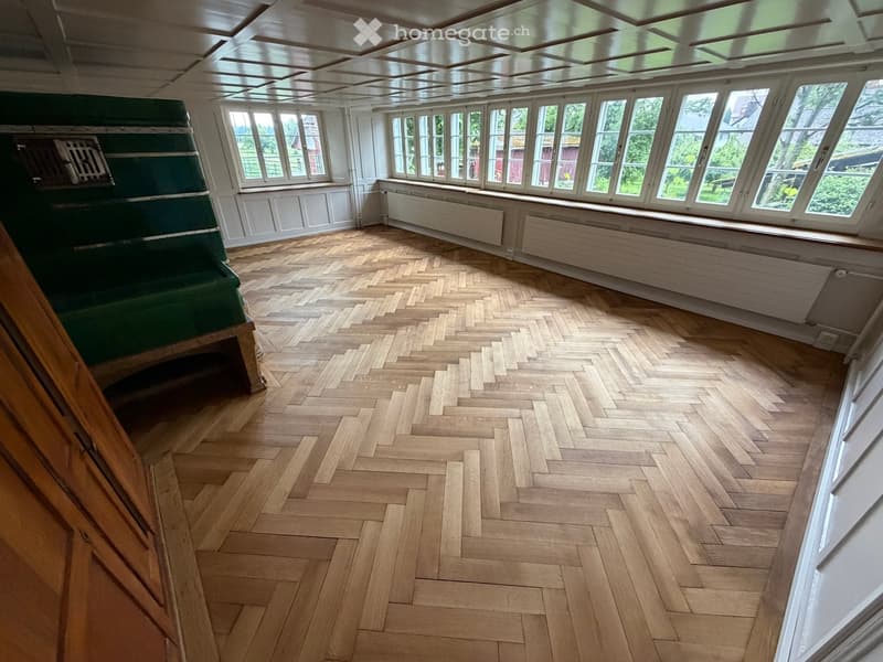 Empty room with herringbone wooden flooring, large windows, white walls, and radiators along the walls.