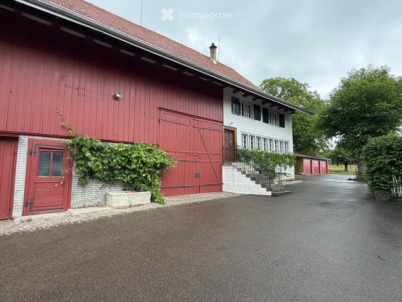 A two-story building with a red barn and a white house connected, featuring a staircase leading to the house entrance, surrounded by greenery and a paved driveway.