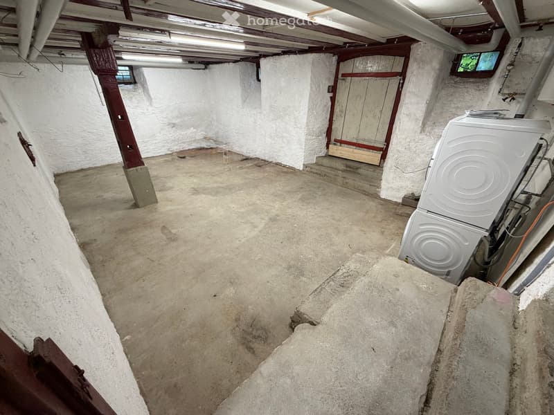 Empty basement with concrete floor, white walls, exposed wooden beams, and a washing machine in the corner.