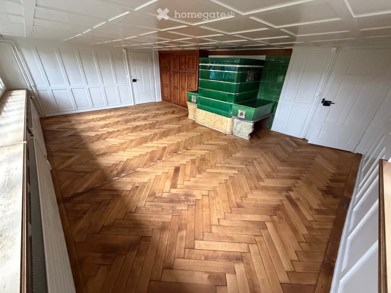 Empty room with herringbone wooden flooring, white paneled walls, and a green fireplace with a stone base.