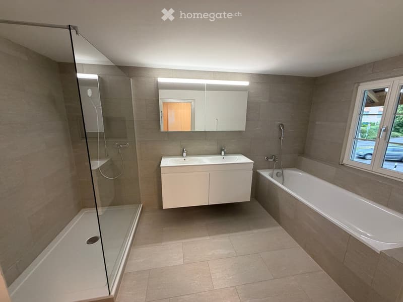 Modern bathroom with a bathtub, double sink, large mirror, and glass-enclosed shower. Light-colored tiles and a window provide natural light.
