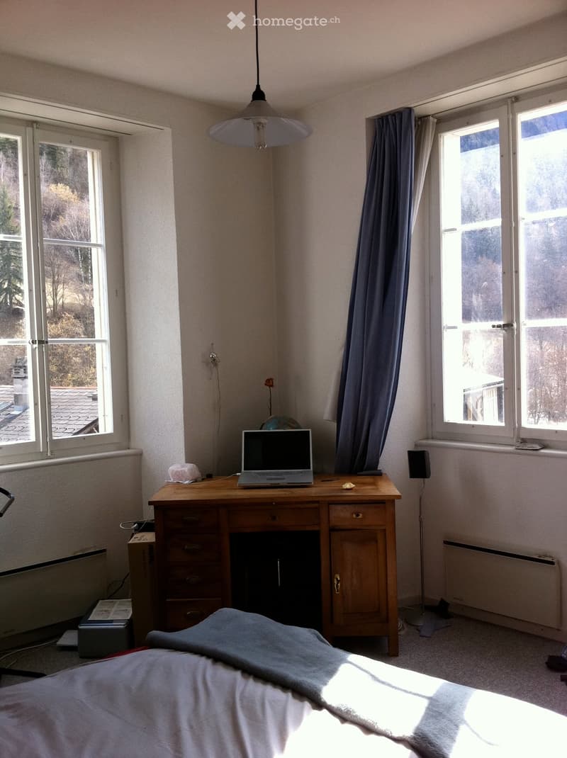 Bright bedroom with a bed, desk with a laptop, globe, and a curtained window. The room has large windows and a radiator.