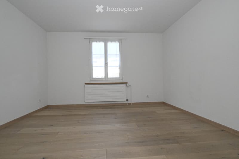 Empty room with wooden flooring, white walls, and a window with a radiator below it.