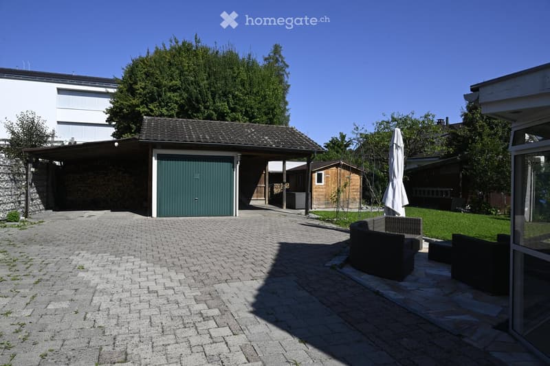 Garden area with a paved brick pathway, outdoor seating area with a sofa and umbrella, garage, and surrounding greenery.