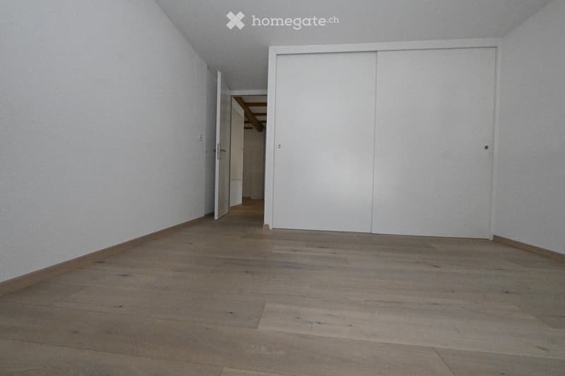 Empty room with wooden flooring and white walls, featuring a closed white sliding door and an open doorway leading to another room.