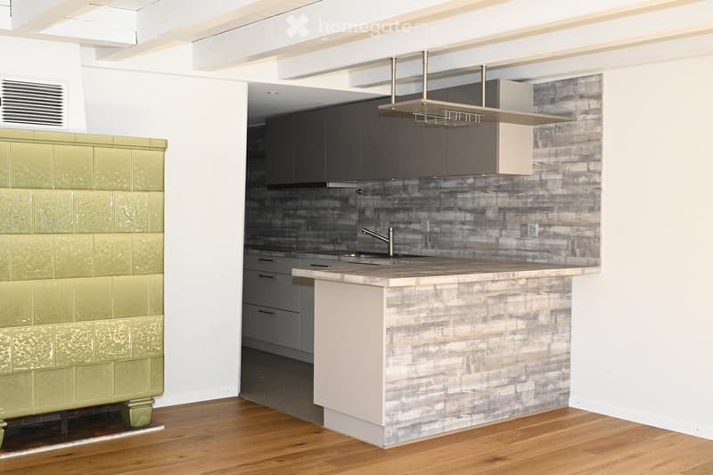 Modern kitchen with a central island, stainless steel sink, dark cabinets, and a range hood. Green tiled backsplash and wooden flooring.