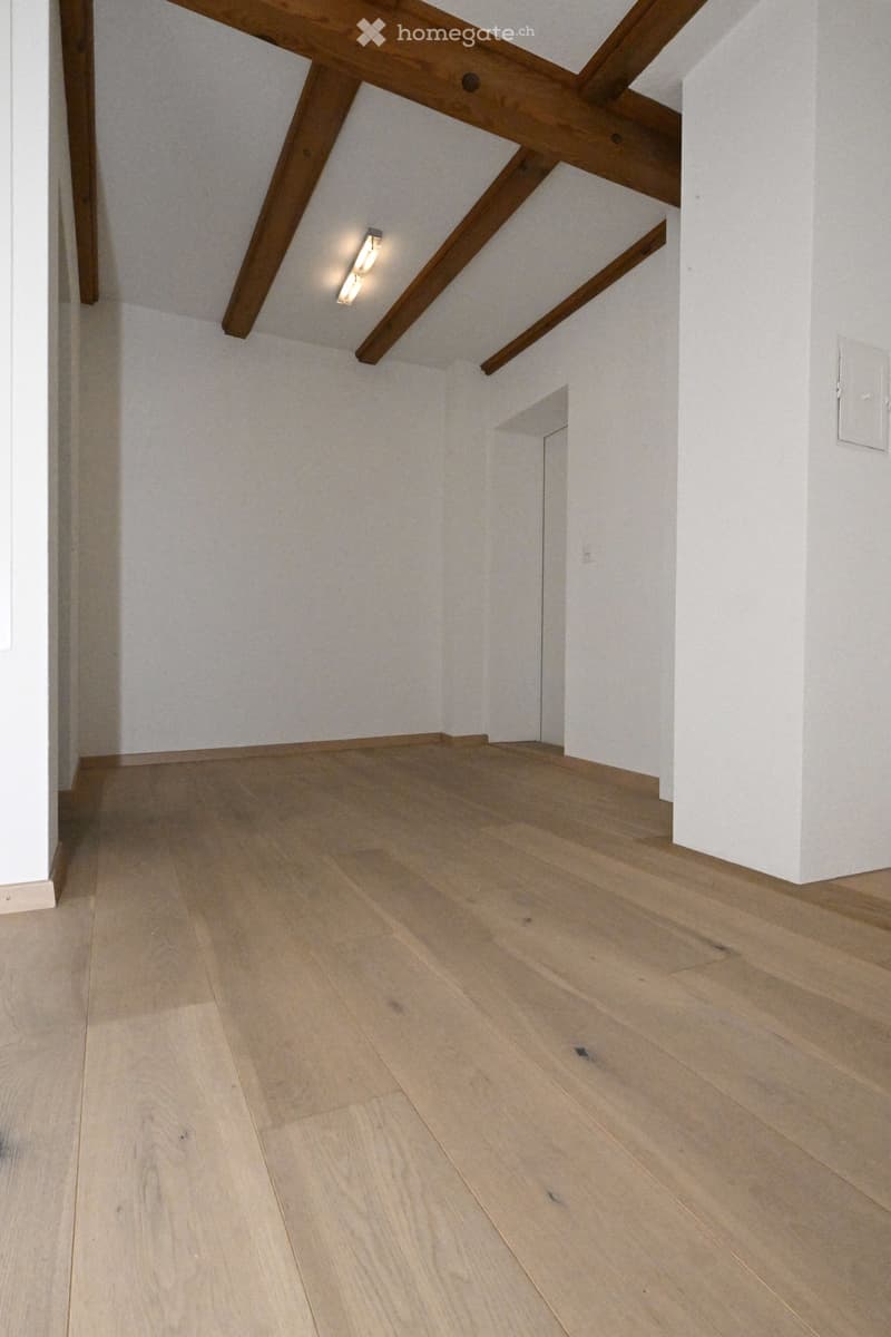 Empty room with light wooden flooring and white walls, featuring exposed wooden beams on the ceiling and a small recessed light fixture.