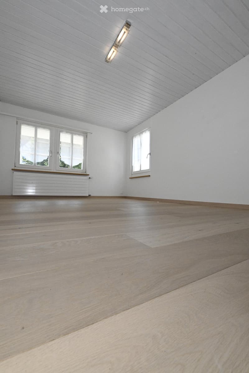 Empty room with light wooden flooring, white walls, and white ceiling with wooden paneling. Two windows with white frames and blinds, and a white radiator under one window.