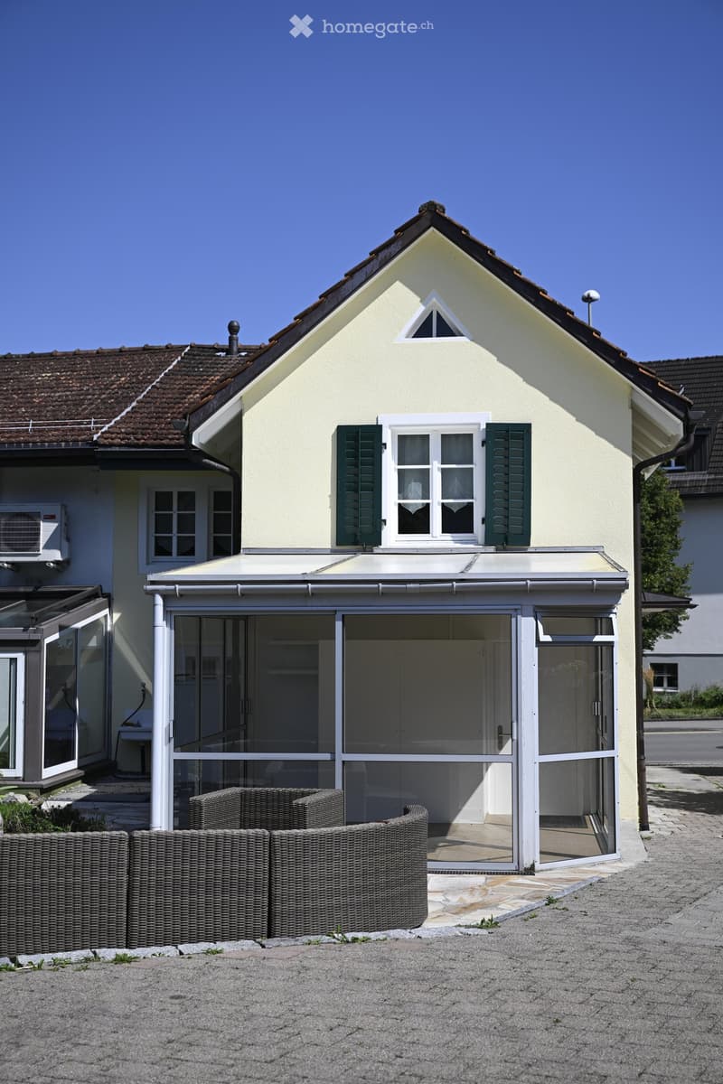 Two-story house with beige exterior, green shutters, and a covered patio area with wicker furniture.