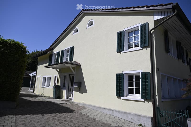 Two-story house with light yellow walls, white-framed windows with green shutters, and a small covered entrance area.