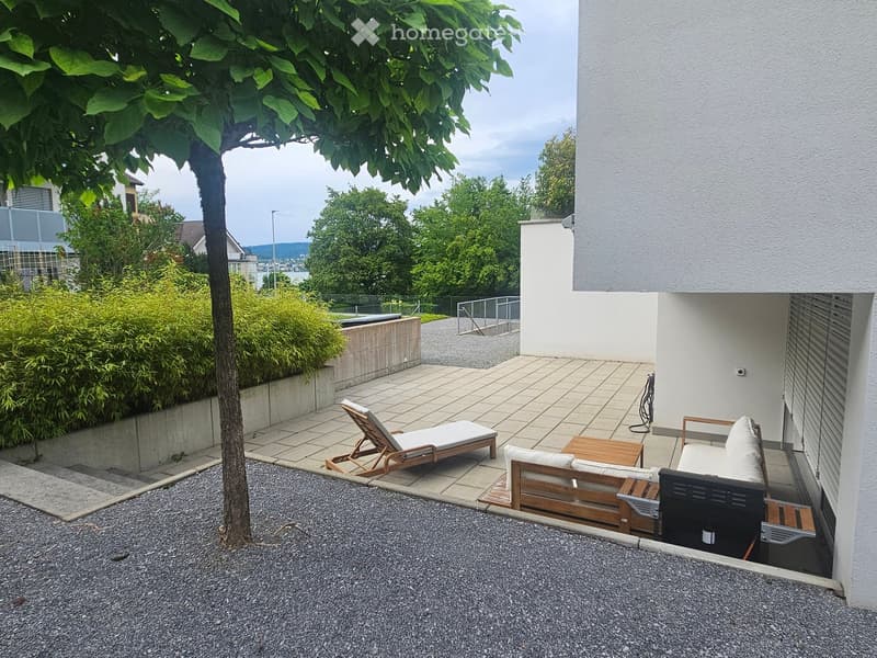 A modern outdoor patio with a tiled floor, a white sofa, a wooden table, a lounge chair, and a tree providing shade. The area is bordered by a low concrete wall and lush greenery.