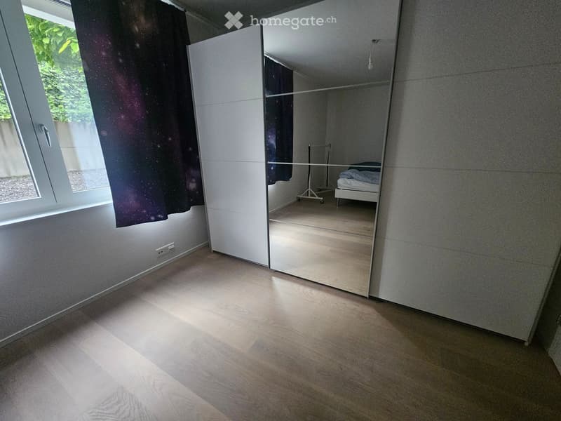 Minimalist bedroom with light wooden flooring, large window, and a single bed visible in the reflection of a full-length mirror.