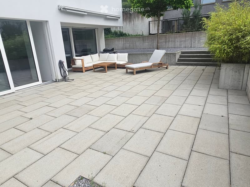 Modern patio with light-colored stone tiles, white sectional sofa, wooden coffee table, and white lounge chair.