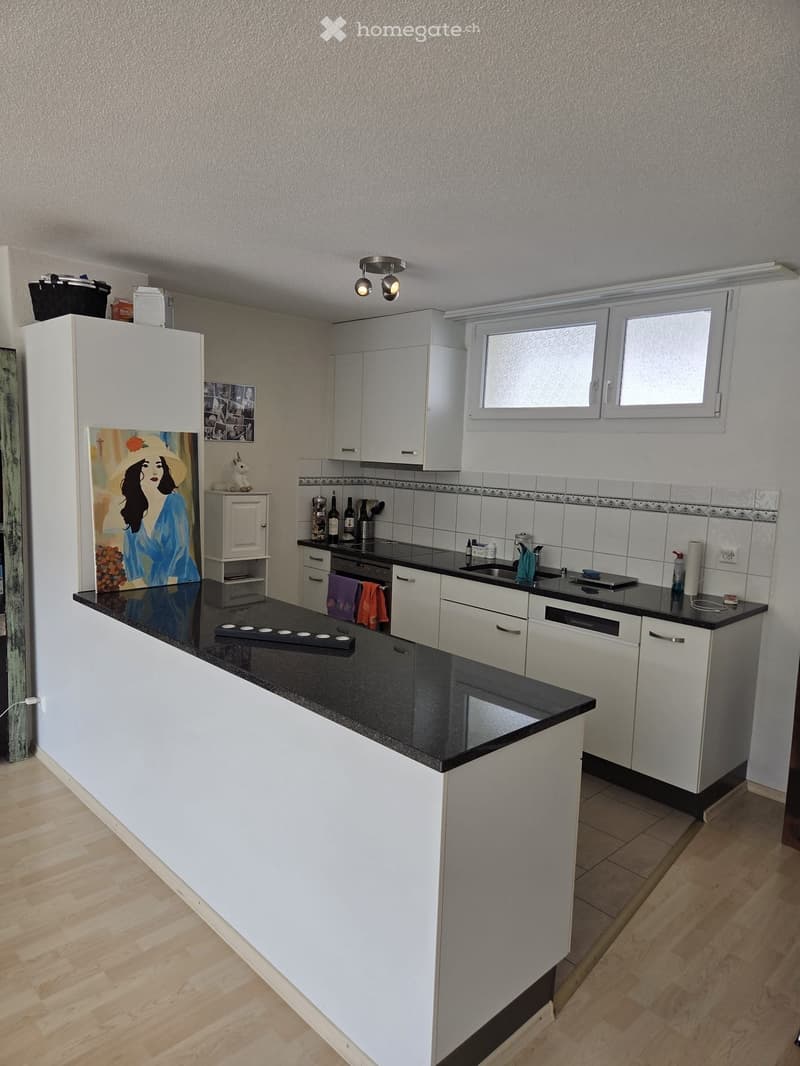 Modern kitchen with white cabinets, black countertop, stainless steel appliances, and a central island. Features include a sink, oven, refrigerator, and decorative items such as a painting and framed photos.