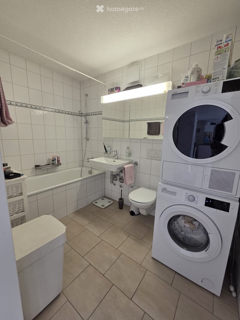 Modern bathroom with white tiles, bathtub, sink, toilet, and a stacked washing machine/dryer unit. Includes a mirror with built-in lighting, towel rack, and cleaning supplies on the washing machine.