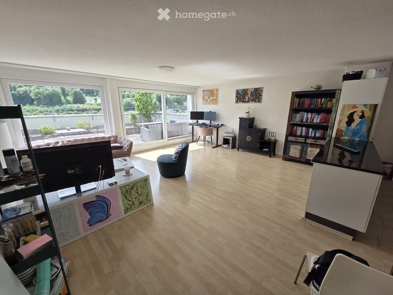 A modern living room with wooden flooring, large windows, a sofa, a chair, a table with a screen, a bookshelf, a stool, a cabinet, and a laptop.