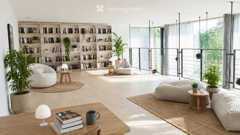 Modern living room with large windows, wooden floor, white walls, and a bookshelf filled with books and plants. Features a wooden table with a lamp, books, and a cup, along with several bean bags and small wooden stools.
