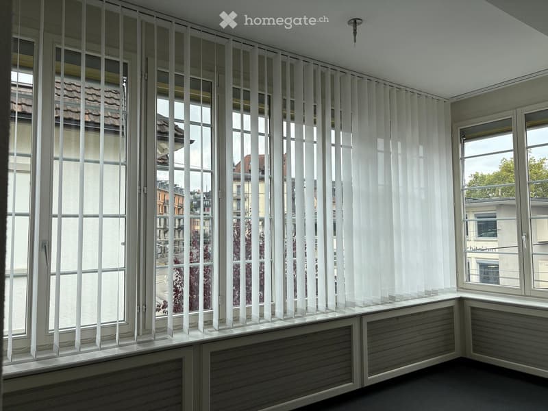 Empty room with large windows covered by vertical blinds, radiator under the windows, and a view of the cityscape.