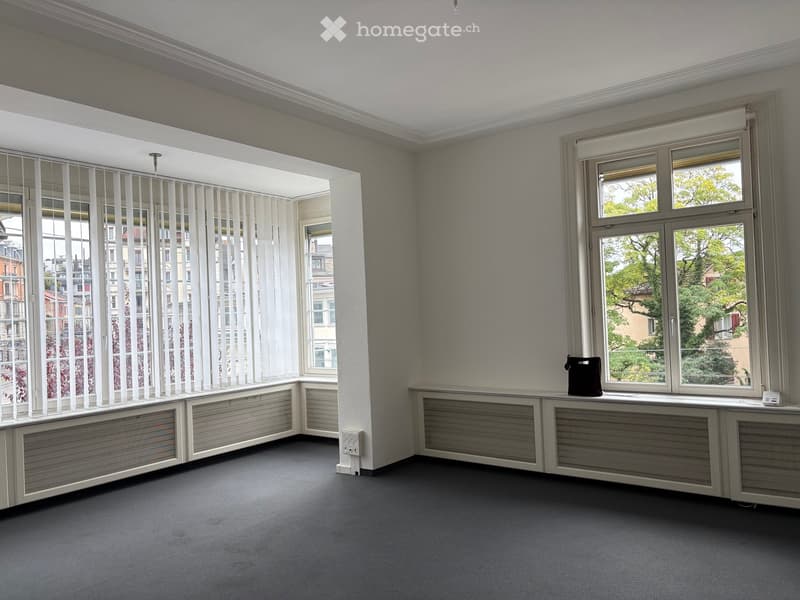 Empty room with grey carpeted floor, white walls, large windows with blinds, radiators, and a small black bag on the windowsill.