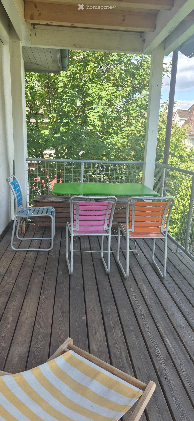 Wooden deck with colorful chairs and a green table, surrounded by a metal railing and lush greenery.