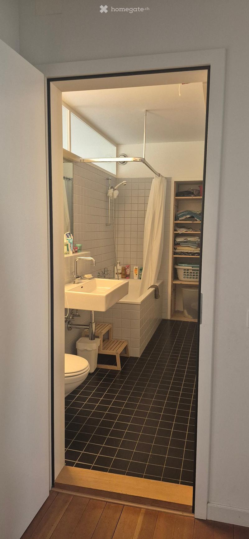 Bathroom with black tiled floor, white sink, toilet, bathtub, shower, and a small wooden shelf holding folded towels and laundry basket.