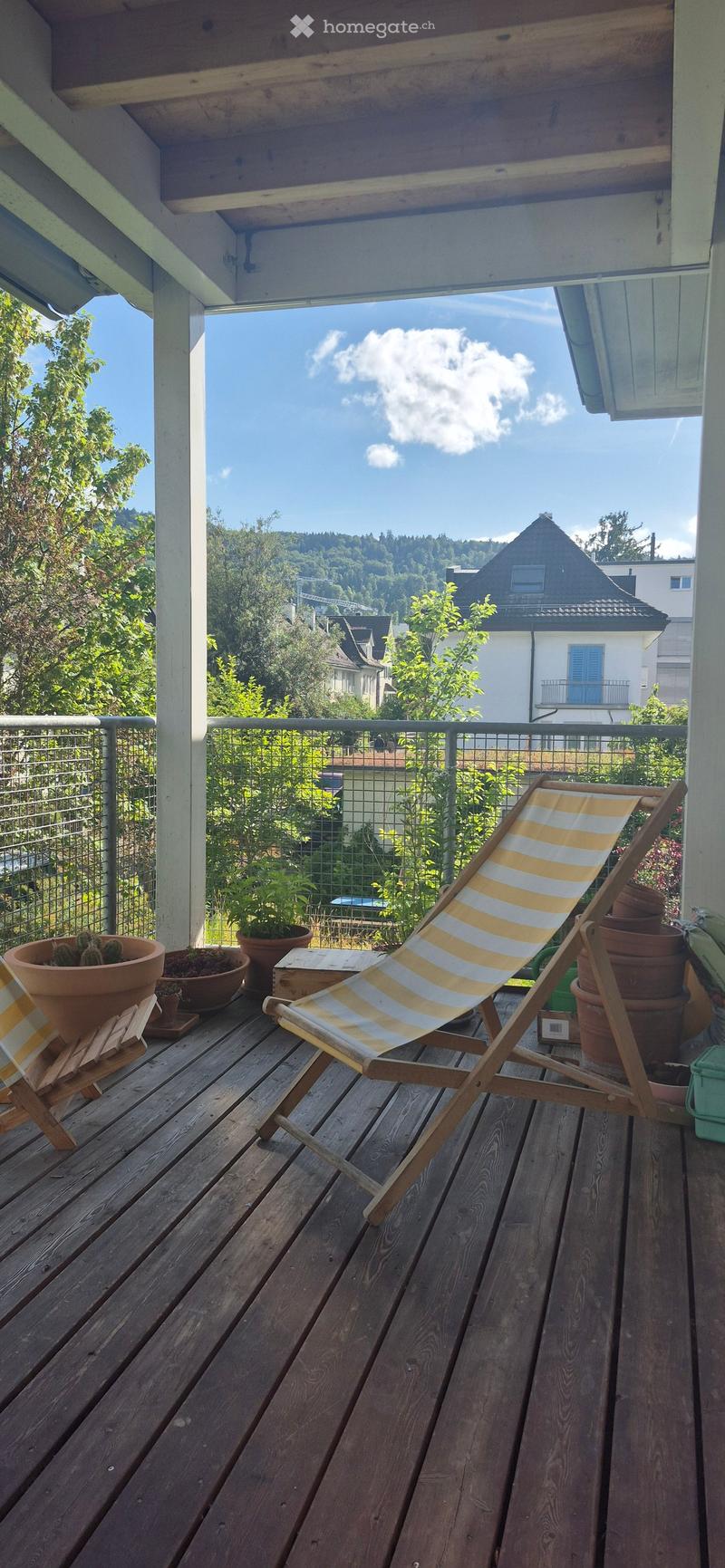 Wooden balcony with a striped beach chair, several potted plants, and a view of a green, hilly landscape.