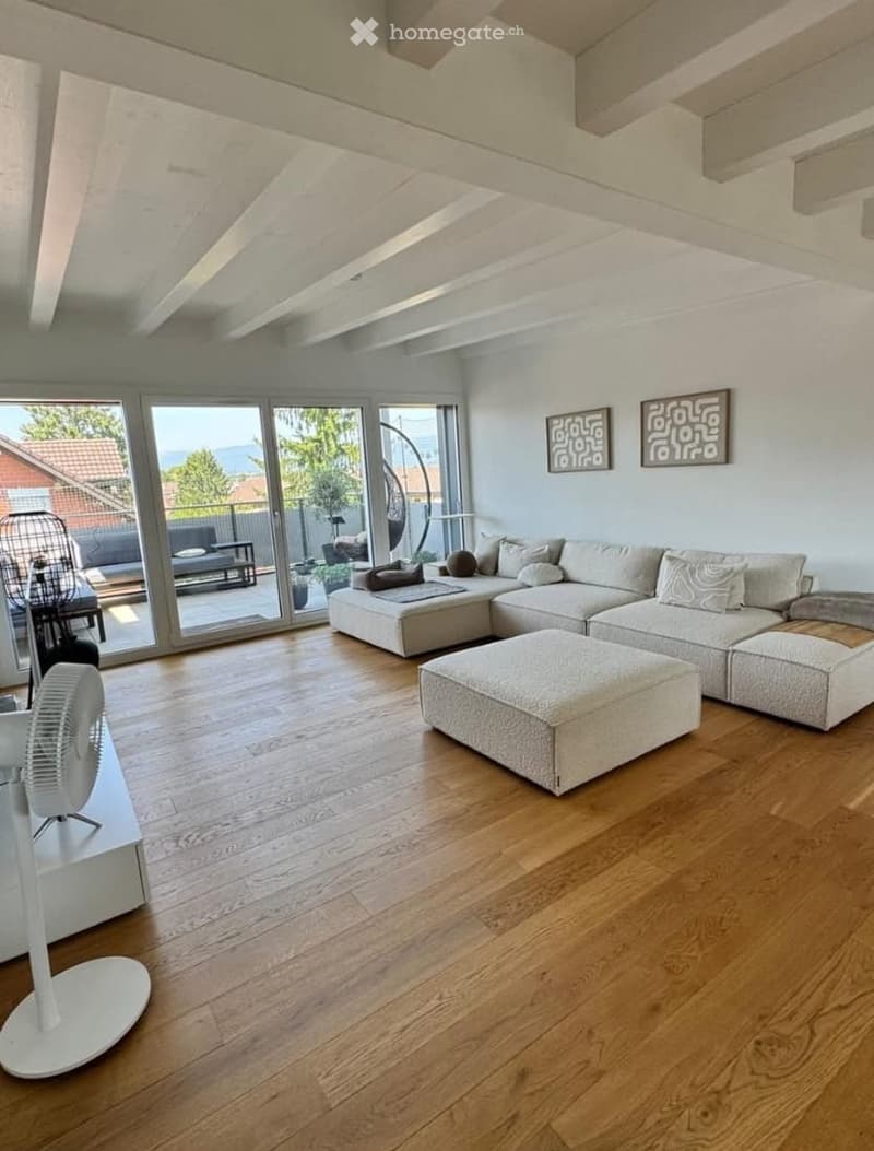 Modern living room with light-colored sectional sofa, ottoman, and a white fan. Large windows offer a scenic view of the outdoors.
