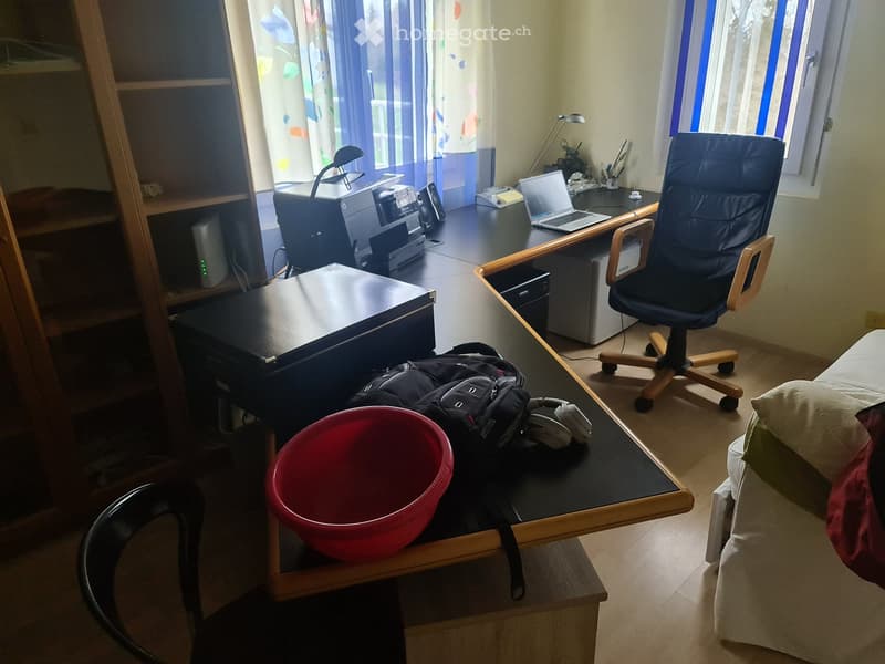 A furnished room with a desk, chair, laptop, printer, sofa, and various personal items including a backpack and headphones.