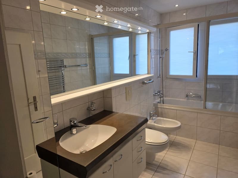 Modern bathroom with a large mirror, a sink with a faucet, a toilet, a bidet, a bathtub, and a shower area. The room features tiled walls and floor, and has a towel rack visible.