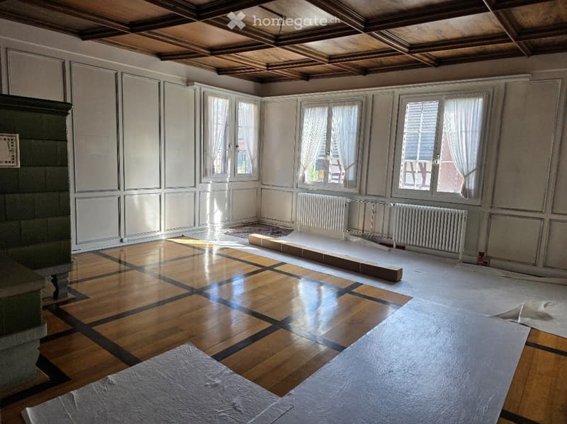 Empty room with wooden flooring, large windows with white curtains, and a fireplace on the left side.