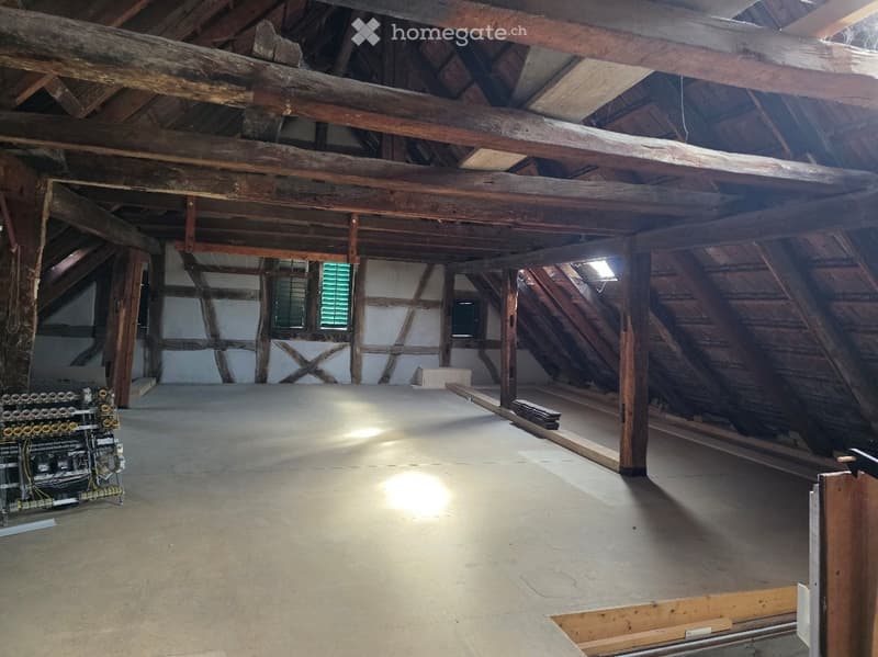 Empty attic space with exposed wooden beams and white plaster walls, featuring small windows and a concrete floor.