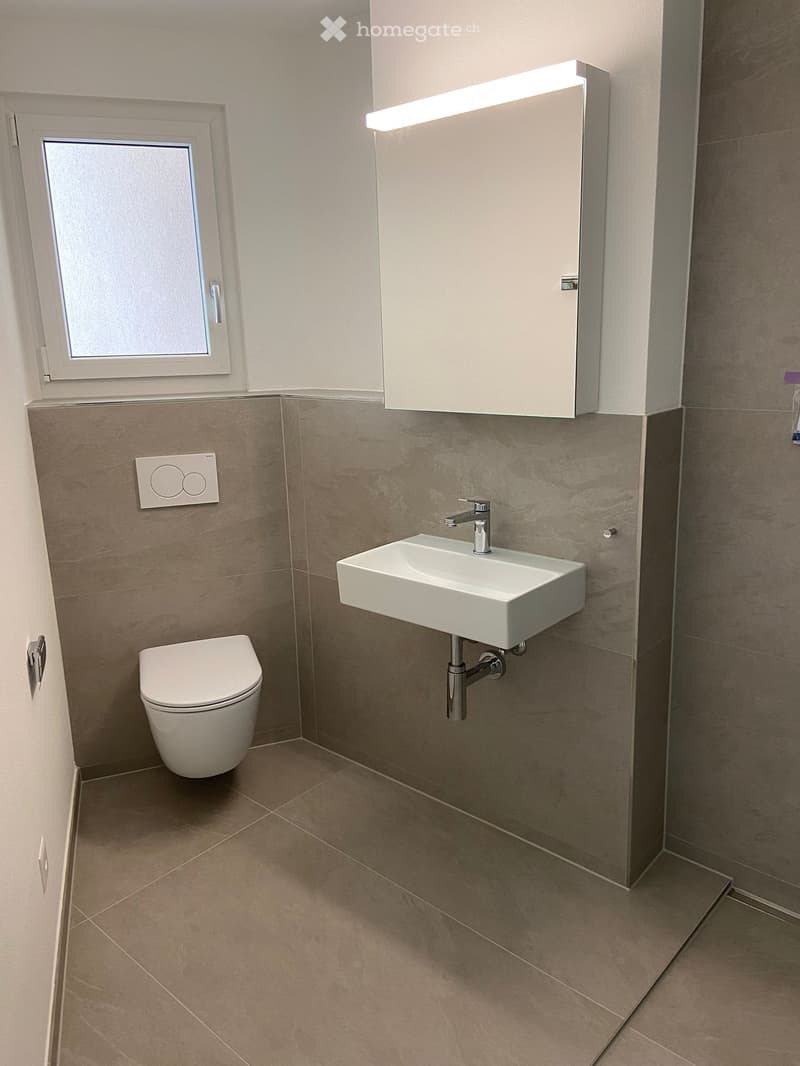 Modern bathroom with a toilet, wall-mounted sink, chrome faucet, and mirrored cabinet with built-in lighting. The walls are tiled in a neutral color scheme, and the floor is covered with beige tiles.