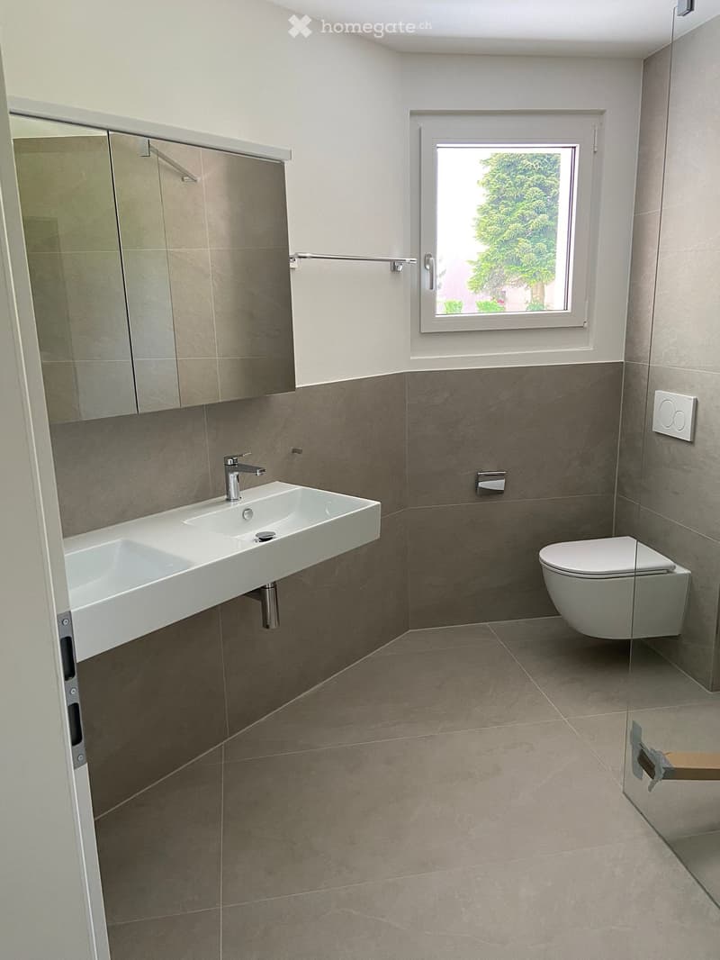 Modern bathroom with a white sink, chrome faucet, toilet, mirrored cabinet, and a window offering a view of greenery outside.