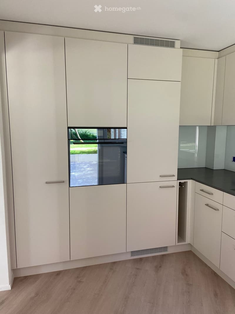 Modern kitchen with white cabinets, a dark countertop, and a built-in oven with a glass door. The floor is made of light wood.