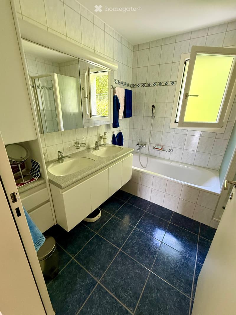 Bathroom with double sink, bathtub, mirror, towels, and a small stool. Blue tiles on the floor and white tiles on the walls.