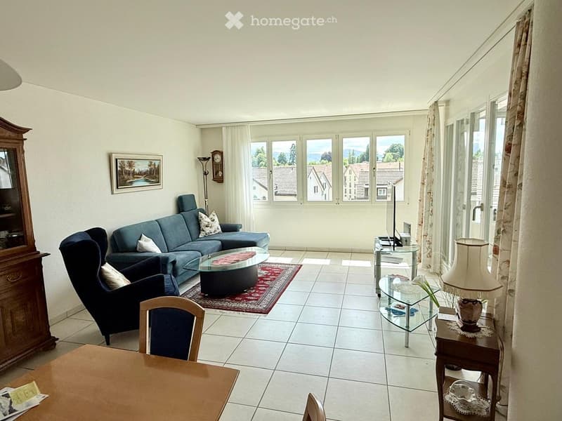 Bright living room with tiled flooring, large windows offering a scenic view, a blue sectional sofa, armchair, coffee table, side table with lamp, and a wooden cabinet.